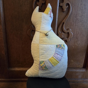 May include: A handmade, quilted cat-shaped pillow in cream and yellow. The cat has a gold bell collar and patchwork details in pastel colors. The pillow is sitting on a black surface against a wooden background.