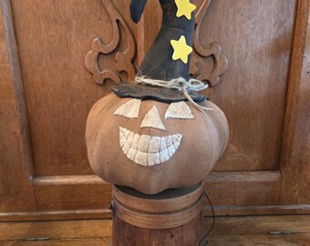 May include: A brown fabric pumpkin with a stitched-on smiling face wearing a black witch hat with yellow stars. The pumpkin is sitting on a wooden bucket.