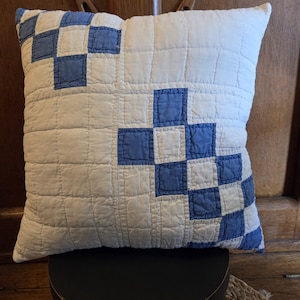 May include: A square throw pillow featuring a white quilted fabric base with a blue patchwork pattern. The design consists of small blue squares arranged in a diagonal, stepped formation across the surface, creating a grid-like texture.
