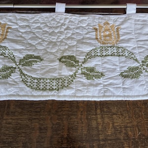 May include: White quilted valance with embroidered floral and leaf design in shades of green and gold. The valance has a rod pocket and white tabs for hanging. The fabric is slightly wrinkled.