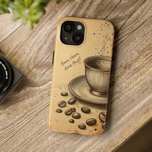 May include: A beige phone case with a vintage illustration of a cup of coffee and coffee beans. The text "Bean there, done that!" is printed on the case.