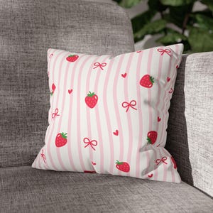 May include: A square decorative pillow with a white background and pink vertical stripes. The pillow is patterned with red strawberries, bows, and hearts. The pillow is placed on a gray sofa.