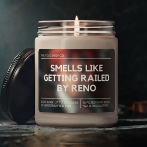 May include: A lit candle in a glass jar with a black lid. The label reads "SMELLS LIKE GETTING RAILED BY RENO." The text "SLOW BURN UP TO 60 HOURS OF UNINTERRUPTED FUN" is also visible. The candle is on a dark surface.
