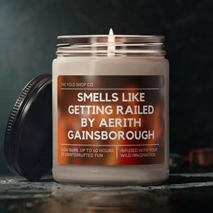 May include: A white scented candle in a glass jar with a black lid. The label reads "SMELLS LIKE GETTING RAILED BY AERITH GAINSBOROUGH". The candle is from "THE YOLD SHOP CO" and promises up to 60 hours of burn time.