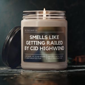 May include: A lit candle in a glass jar with a black lid. The label reads "SMELLS LIKE GETTING RAILED BY CID HIGHWIND." The candle is from The Yold Shop Co. and promises up to 60 hours of burn time.