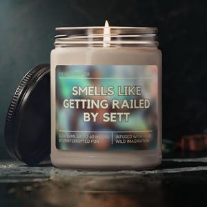 May include: A lit candle in a glass jar with a black lid. The label reads "SMELLS LIKE GETTING RAILED BY SETT". The candle is white with a silver rim. The text on the label also says "LOW BURN: UP TO 60 HOURS OF UNINTERRUPTED FUN" and "INFUSED WITH YOUR WILD IMAGINATION".