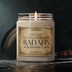 May include: A lit soy wax candle in a glass jar with a silver lid. The label reads "SMELLS LIKE GETTING RAILED BY RADAHN at Redmane Castle." The candle is 9 oz. and from The YOLO Shop Co.