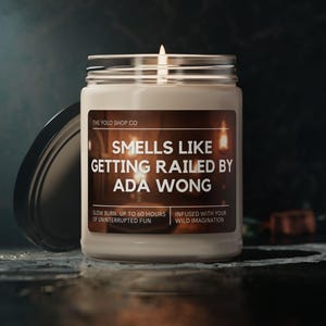 May include: A lit white candle in a clear glass jar with a silver lid. The label reads "SMELLS LIKE GETTING RAILED BY ADA WONG". The candle is from "THE YOLD SHOP CO" and promises up to 60 hours of burn time.