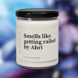 Peut inclure: Bougie en cire de soja blanche dans un pot en verre transparent avec un couvercle noir. L'étiquette indique "Smells like getting railed by Ahri" et "9 oz Soy Wax Candle". La marque "The Yolo Shop Co" est également imprimée.