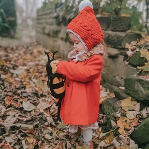 May include: A child in a vibrant red coat and a crocheted red hat with a white bobble. The child is holding a black and yellow stuffed bee toy. The scene is set amongst fallen leaves and a stone wall.
