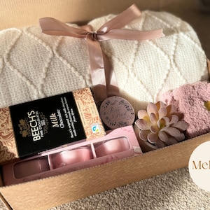 Pu&ograve; includere: Una scatola regalo di cartone marrone piena di una coperta di maglia bianca, una coperta pelosa rosa, una piccola succulenta in vaso, una scatola di cioccolato, una piccola candela e un piccolo contenitore rotondo con il testo "Rose Petal" su di esso. La scatola &egrave; etichettata "Mehugs".
