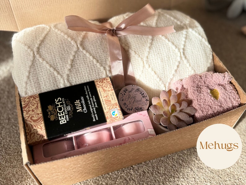 Pu&ograve; includere: Una scatola regalo di cartone marrone piena di una coperta di maglia bianca, una coperta pelosa rosa, una piccola succulenta in vaso, una scatola di cioccolato, una piccola candela e un piccolo contenitore rotondo con il testo "Rose Petal" su di esso. La scatola &egrave; etichettata "Mehugs".