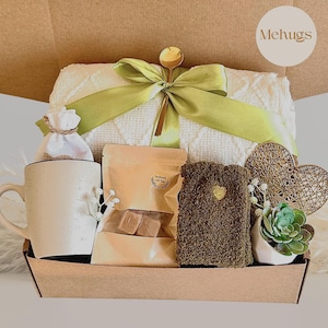 Può includere: Una confezione regalo piena di una coperta bianca, una tazza, un sacchetto di tè, prodotti da bagno, calzini, un cuore decorativo e una piccola pianta succulenta. La scatola è legata con un nastro verde e include un cucchiaio d'oro.
