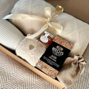 May include: A gift box filled with cosy items, including a white mug, a soft blanket, a small jar of honey, a bar of milk chocolate, and a small hessian bag. The box is tied with a cream-coloured ribbon.