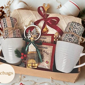 Può includere: Un cofanetto regalo con due tazze bianche, ciascuna con un rotolo di tessuto avvolto. La scatola contiene anche bustine di caffè, un ornamento decorativo e una bottiglietta. Un nastro rosso e un cucchiaio dorato aggiungono tocchi festosi. Il marchio "Mehugs" è visibile.