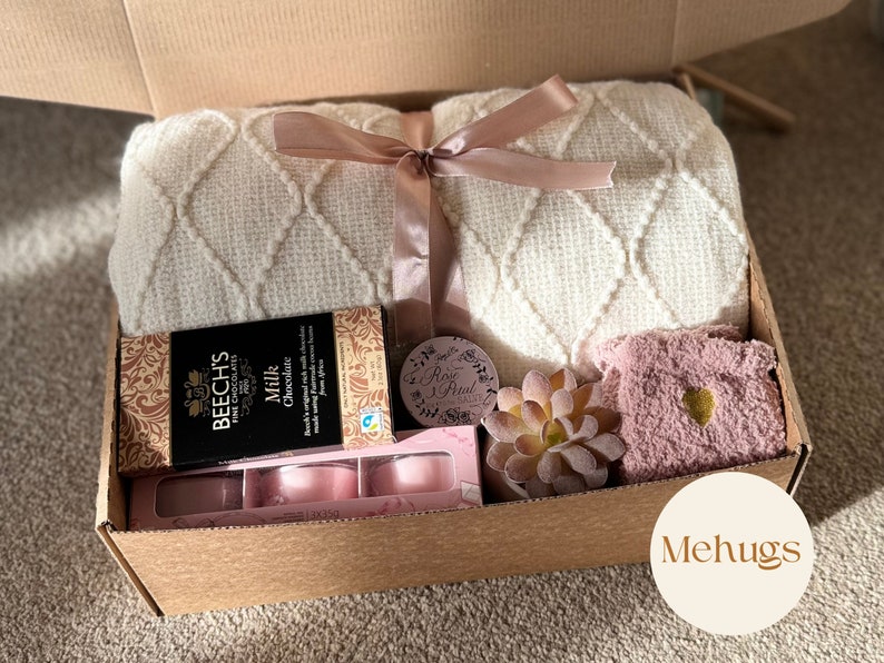 Pu&ograve; includere: Una scatola di cartone marrone contenente una coperta bianca e crema, una scatola di cioccolato, una candela rosa, una piccola pianta grassa e un calzino rosa con un cuore dorato. La scatola &egrave; etichettata "Mehugs".