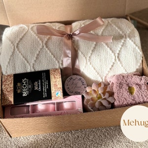 Pu&ograve; includere: Una scatola di cartone marrone con una coperta bianca e crema, una scatola di cioccolato, una candela rosa, una piccola pianta grassa e una ciabatta rosa pelosa. La scatola &egrave; legata con un nastro rosa e ha la scritta "Mehugs" stampata sul lato.