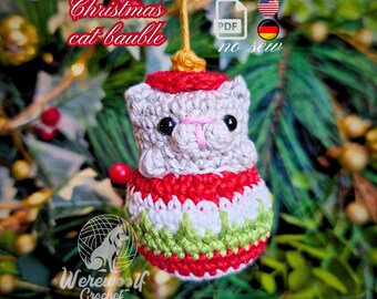 May include: A crocheted Christmas cat bauble ornament featuring a light gray cat head with black eyes and a pink nose, sitting atop a round, red, white, and green patterned base. The ornament hangs from a gold string and includes text.