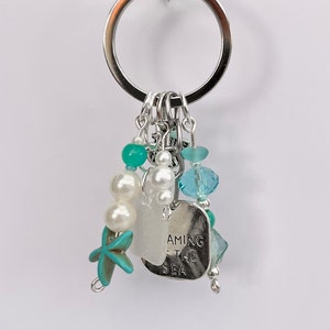 May include: A silver key chain with a charm that says "Dreaming of the Sea". The charm is surrounded by white pearls, turquoise and blue beads, and a turquoise starfish.