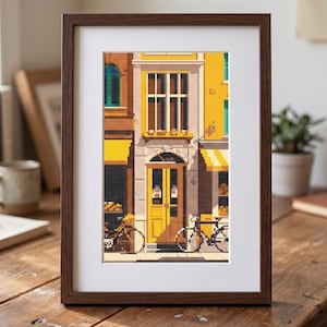 May include: A framed pixel art print depicting a street-level view of a building facade with a yellow door, large windows, and bicycles parked in front. The artwork features a color palette of yellow, brown, and white, encased in a dark wood frame.