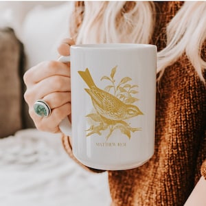 May include: White ceramic mug with a gold bird and foliage design. The mug has the text "Matthew 10:31" printed on the bottom.