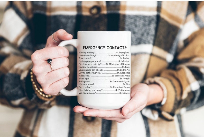 May include: White ceramic mug with black text that reads "EMERGENCY CONTACTS:" followed by a list of common problems and the corresponding saint to call for help.