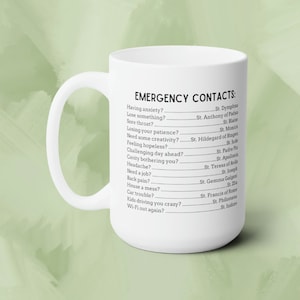 May include: White ceramic coffee mug with a black text design that reads "Emergency Contacts" and lists various situations with a corresponding saint to call upon for help.