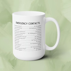 May include: White ceramic mug with black text that reads "EMERGENCY CONTACTS:" and lists a series of humorous situations with corresponding saint names.  The mug has a black handle.