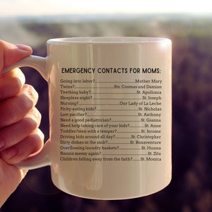 K&ouml;nnte beinhalten: Eine wei&szlig;e Keramiktasse mit schwarzem Henkel und dem Text "EMERGENCY CONTACTS FOR MOMS:" gefolgt von einer Liste g&auml;ngiger Herausforderungen der Elternschaft und dem entsprechenden Heiligen, an den man sich wenden kann.