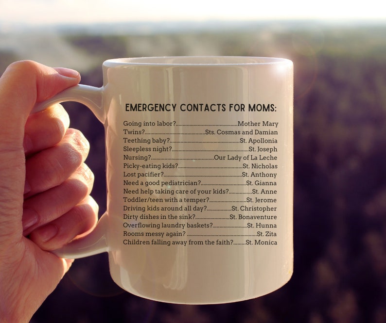 K&ouml;nnte beinhalten: Eine wei&szlig;e Keramiktasse mit schwarzem Henkel und dem Text "EMERGENCY CONTACTS FOR MOMS:" gefolgt von einer Liste g&auml;ngiger Herausforderungen der Elternschaft und dem entsprechenden Heiligen, an den man sich wenden kann.