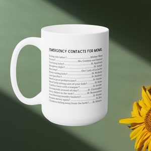 K&ouml;nnte beinhalten: Eine wei&szlig;e Keramik-Kaffeetasse mit einem schwarzen Textdesign, das "Notfallkontakte f&uuml;r M&uuml;tter" lautet, mit einer Liste von h&auml;ufigen Situationen in der Elternschaft und den entsprechenden Heiligen, die man um Hilfe bitten kann. Die Tasse steht auf einer gr&uuml;nen Oberfl&auml;che mit einer gelben Sonnenblume im Hintergrund.