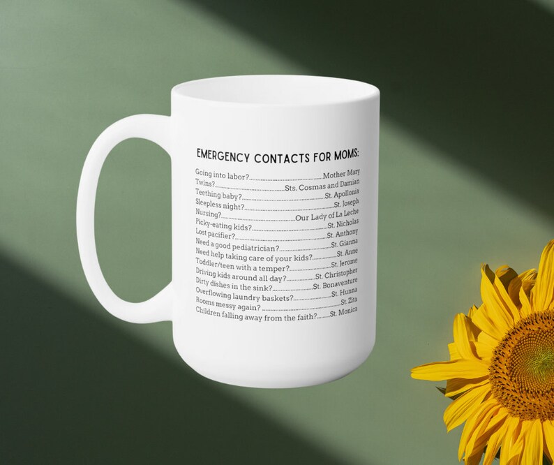 K&ouml;nnte beinhalten: Eine wei&szlig;e Keramik-Kaffeetasse mit einem schwarzen Textdesign, das "Notfallkontakte f&uuml;r M&uuml;tter" lautet, mit einer Liste von h&auml;ufigen Situationen in der Elternschaft und den entsprechenden Heiligen, die man um Hilfe bitten kann. Die Tasse steht auf einer gr&uuml;nen Oberfl&auml;che mit einer gelben Sonnenblume im Hintergrund.