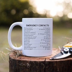 May include: White ceramic mug with a black handle and the text "EMERGENCY CONTACTS:" followed by a list of saints and their corresponding problems. The mug is sitting on a tree stump with a pair of yellow pruning shears.