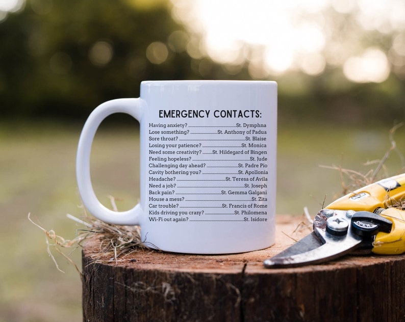 May include: White ceramic mug with a black handle and the text "EMERGENCY CONTACTS:" followed by a list of saints and their corresponding problems. The mug is sitting on a tree stump with a pair of yellow pruning shears.