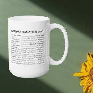 K&ouml;nnte beinhalten: Wei&szlig;e Keramiktasse mit einem schwarzen Textdesign, das "Notfallkontakte f&uuml;r M&uuml;tter" lautet und eine Reihe von g&auml;ngigen Herausforderungen der Elternschaft mit dem Namen eines Heiligen neben jedem auflistet.