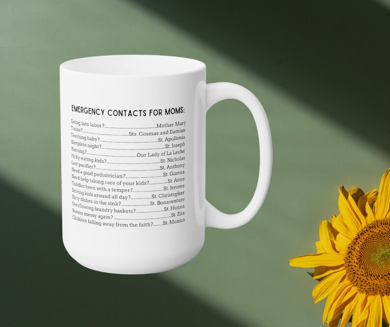 K&ouml;nnte beinhalten: Wei&szlig;e Keramiktasse mit einem schwarzen Textdesign, das "Notfallkontakte f&uuml;r M&uuml;tter" lautet und eine Reihe von g&auml;ngigen Herausforderungen der Elternschaft mit dem Namen eines Heiligen neben jedem auflistet.