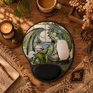 Könnte beinhalten: Ein rundes Mauspad mit einer grünen Drachenillustration im Buntglas-Stil und einer schwarzen Handgelenkauflage. Eine weiße Computermaus liegt auf der Oberfläche des Pads, das komplizierte grüne und weiße Glasmuster zeigt.