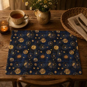 Puede incluir: Un mantel individual rectangular con un fondo azul marino oscuro y un patrón repetitivo de soles, lunas y orbes celestiales dorados. El diseño incluye motivos circulares de varios tamaños con líneas radiantes y texturas.