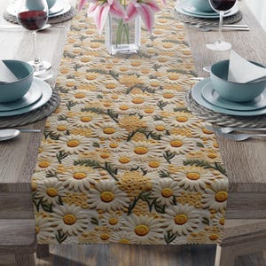 Puede incluir: Camino de mesa beige con un estampado de margaritas en 3D, sobre una mesa de madera. El camino de mesa presenta pétalos de margarita blancos con centros amarillos y hojas verdes. La mesa está puesta con platos, cuencos, cubiertos y copas de vino.