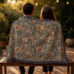 May include: A woven throw blanket featuring a dense, intricate floral pattern in shades of muted gold, cream, and deep navy blue. The rectangular textile has a fringed edge along the bottom and sides, displaying a detailed botanical design.
