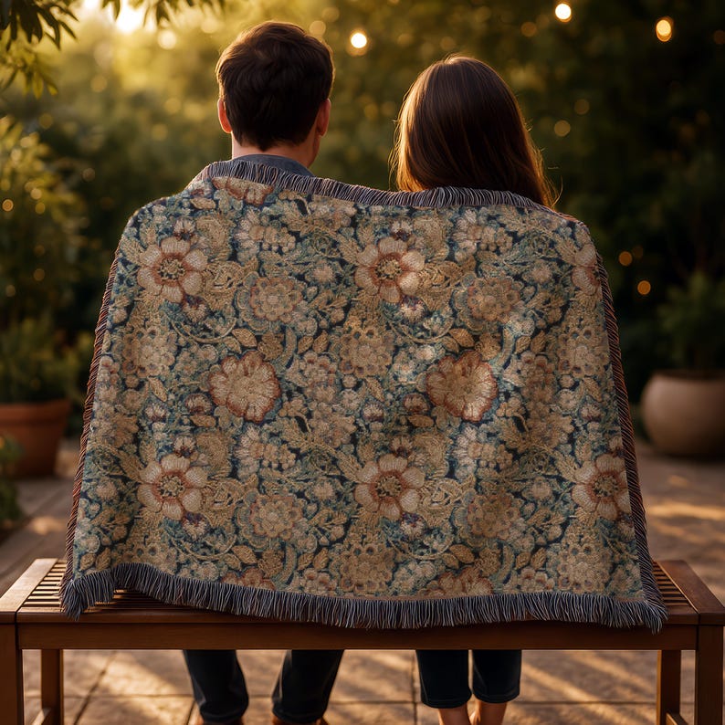 May include: A woven throw blanket featuring a dense, intricate floral pattern in shades of muted gold, cream, and deep navy blue. The rectangular textile has a fringed edge along the bottom and sides, displaying a detailed botanical design.