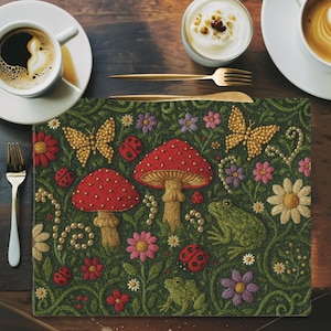 Op de afbeelding: Een decoratieve placemat met een grillig boslandschap. Het ontwerp omvat rode paddenstoelen, gouden vlinders, lieveheersbeestjes en groene kikkers, tegen een donkergroene achtergrond met geborduurde bloemen en wijnranken. De placemat is rechthoekig.