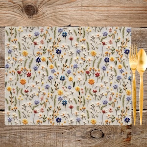 May include: A white placemat with a floral pattern embroidered in shades of blue, yellow, red, and green. The placemat is made of a soft, textured fabric and features a variety of small flowers and leaves.
