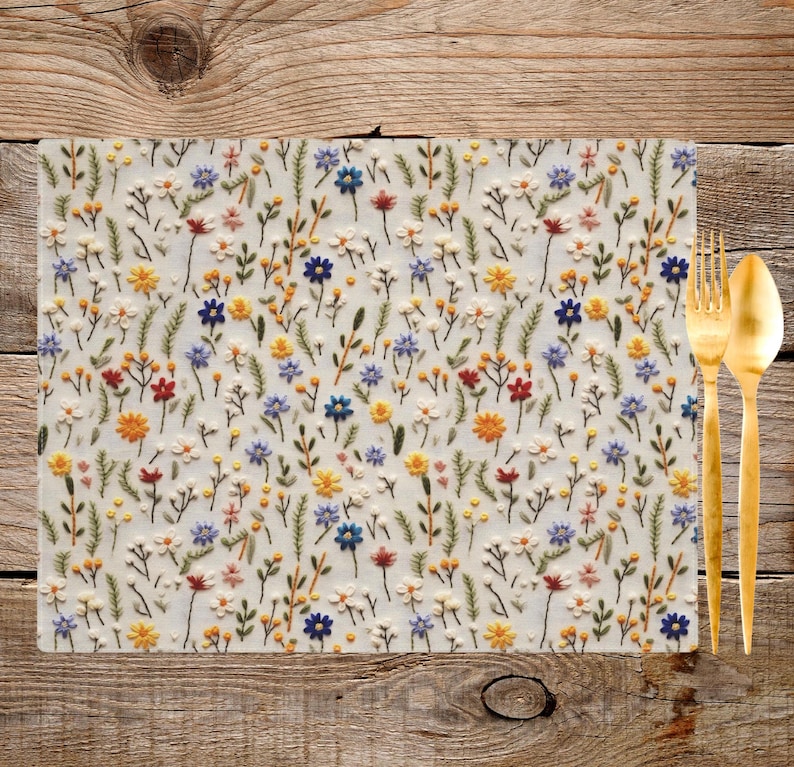 May include: A white placemat with a floral pattern embroidered in shades of blue, yellow, red, and green. The placemat is made of a soft, textured fabric and features a variety of small flowers and leaves.