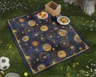 May include: A rectangular picnic blanket featuring a dark blue background with a repeating gold celestial pattern of suns, moons, and stars. The blanket has a thick gold border and is displayed on grass with a small basket and food items.