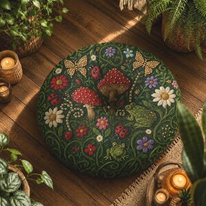 Puede incluir: Un cojín de suelo redondo de color verde oscuro con bordados detallados de setas rojas, flores blancas y moradas, dos mariposas doradas, mariquitas y dos ranas verdes, todo ello rodeado de un diseño de enredaderas entrelazadas.