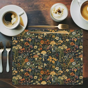 May include: A dark placemat with a botanical design featuring mushrooms, flowers, and foliage in shades of orange, yellow, and green. The placemat is rectangular and sits on a dark wooden table, alongside silverware, coffee, and a small bowl of cream.
