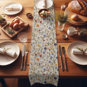 May include: A white table runner with a floral embroidery design. The embroidery features small, colorful flowers and leaves in shades of red, yellow, blue, and green. The runner is laid out on a wooden table with place settings.