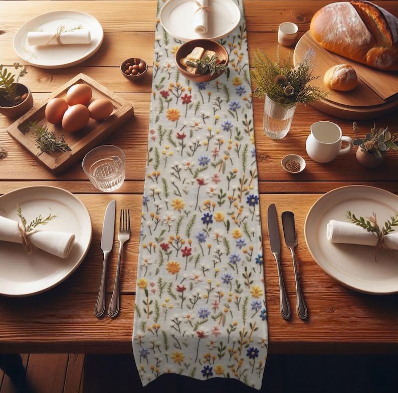 May include: A white table runner with a floral embroidery design. The embroidery features small, colorful flowers and leaves in shades of red, yellow, blue, and green. The runner is laid out on a wooden table with place settings.