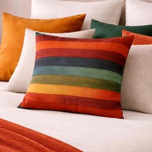 May include: A collection of decorative pillows in various colors on a bed. The pillows include solid orange, white, green, and a striped pillow with bands of red, orange, yellow, green, and blue. The bed has a white comforter and an orange throw.